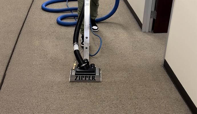 A professional using a Zipper brand carpet cleaning wand and vacuum hose system to deep clean a neutral-toned commercial carpet in a hallway