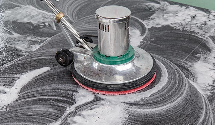 A close-up of a professional floor scrubbing machine with a red rotating pad cleaning a dark grey tiled surface