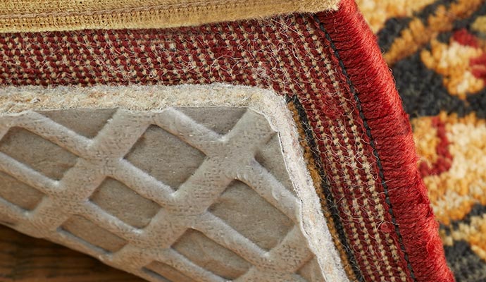 Close-up of a decorative red and gold area rug pulled back to reveal a grey rubberized non-slip grid rug pad underneath