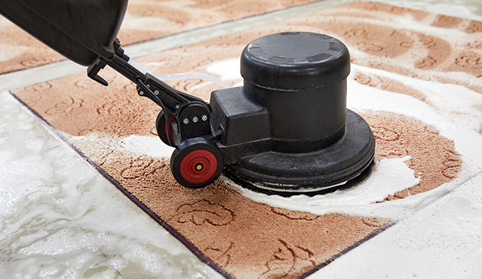 A technician using a rotary floor machine to deeply shampoo and clean a patterned beige area rug.