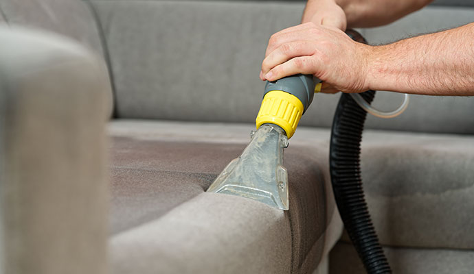 Close-up of a technician using a specialized steam extraction hand tool to deep clean a grey fabric sofa.