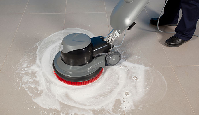 A technician using a heavy-duty rotary floor machine with a red brush to deep clean beige floor tiles and grout lines.