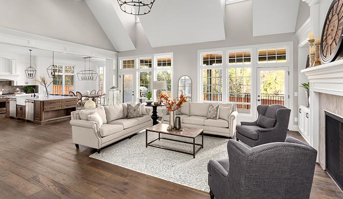 A spacious open-concept great room featuring a grand living area with beige sofas, charcoal wingback chairs, and a fireplace