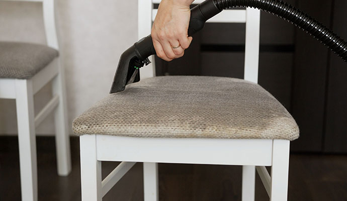 Close-up of a hand using a professional vacuum extraction tool to clean a gray dining chair seat.