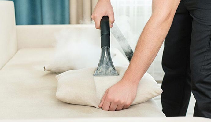 A close-up of a professional cleaner using a steam extraction tool to deep clean a beige fabric sofa pillow